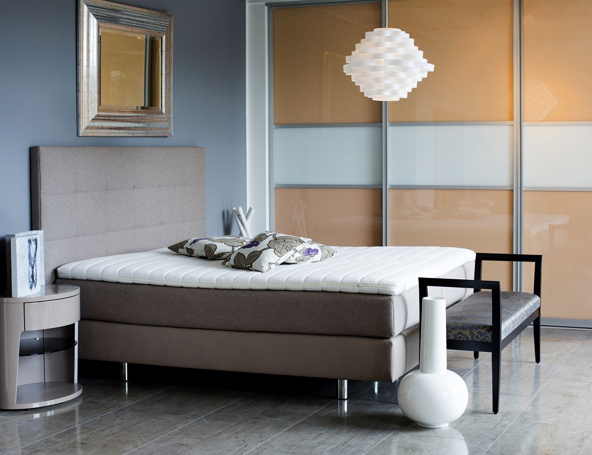 Stilvolles Schlafzimmer mit einem modernen Bett mit dekorativen Kissen, einer runden Pendelleuchte und minimalistischen Möbeln vor grauem Hintergrund.