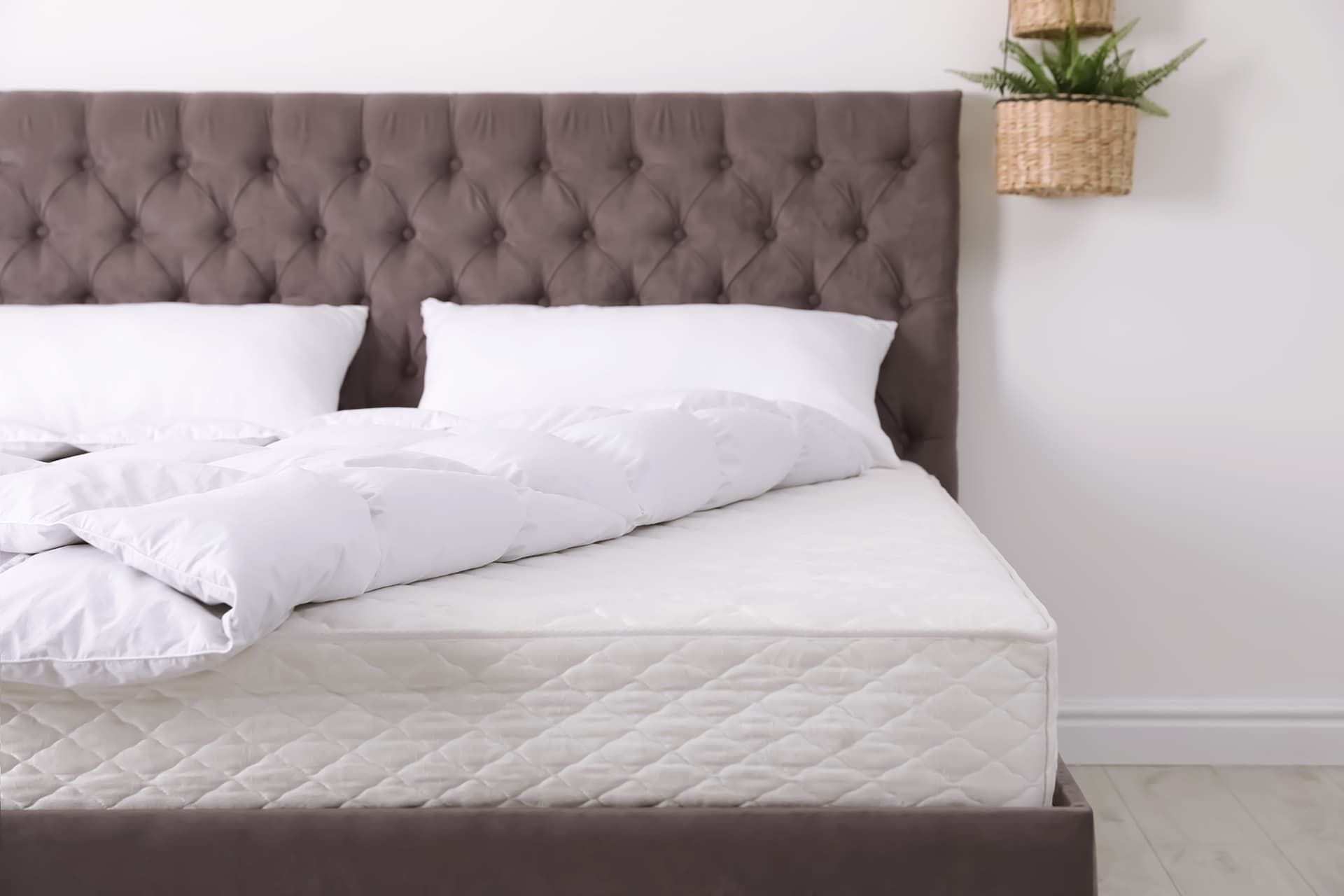 Ein weiches, getuftetes graues Kopfteil thront über einem ordentlich gemachten Bett mit weißen Kissen und einer flauschigen weißen Bettdecke auf einem hellen Holzboden.